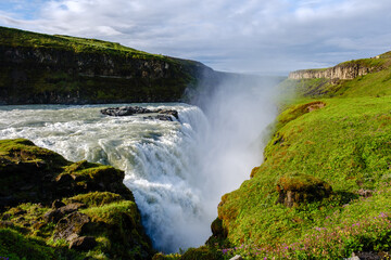 Fototapeta premium Water flows powerfully over the Gullfoss waterfall in Iceland, creating mist that dances with sunlight.