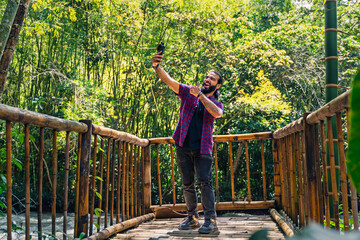 Man in plaid shirt with tied hair photographing himself on wooden bridge in lush Colombian jungle...