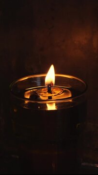 Vertical video close-up of a flickering flame in an oil lamp inside a church in Elefsina, Greece. The flame reflects softly in the glass holder. Background is dark. No people.