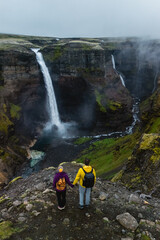 Two explorers stand hand in hand on a rocky ledge, gazing at breathtaking Haifoss waterfalls...