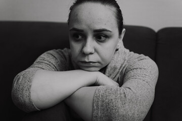 A woman sits on a couch hugging her knees and gazing off into the distance with a thoughtful...