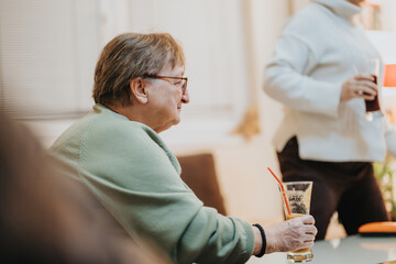 Elderly woman holding a drink seated at a social gathering indoors, sharing moments with others in...