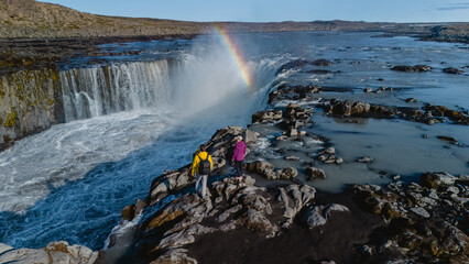 Two adventurers stand on rocky terrain near Dettifoss, observing the majestic Selfoss waterfall...