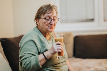 A senior woman relaxing at home, sipping a nutritious fruit smoothie through a straw in a...