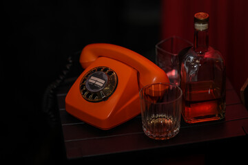 Vintage orange rotary phone, glass of whiskey, and liquor bottle on a dark wooden table in moody...