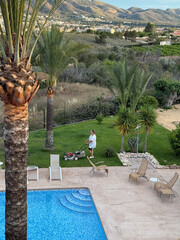 Individual mowing lush green lawn near swimming pool, surrounded by palm trees and scenic...