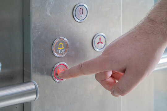 Human hand activates the illuminated red emergency stop button on an modern elevator panel, indicating immediate safety action and urgent crisis intervention