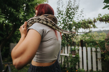 A girl with red hair cradles a snake on her shoulders in a vibrant garden. The scene portrays...