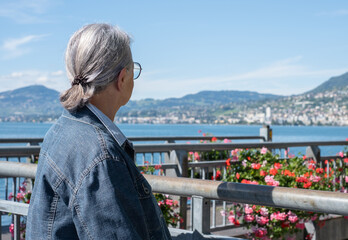 Fototapeta premium Rear view of a gray-haired senior woman sitting in front of the Geneva lake admiring the landscape, a relaxed senior grandmother enjoying peaceful moments in retirement