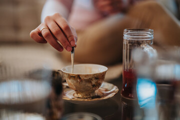 Close-up of a hand stirring tea in an elegant floral teacup, surrounded by a cozy atmosphere with glasses and warm hues, evoking a feeling of relaxation and calmness.