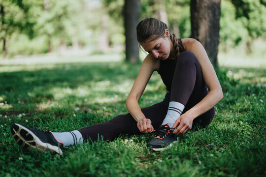 A young woman in athletic attire preparing for exercise in a serene, grassy park setting. She is tying her shoelaces while enjoying the peaceful ambiance of the outdoors. - Powered by Adobe
