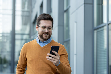 Close-up photo of a young man standing on the street and smiling while using his mobile phone