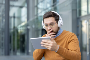 Close-up photo of a young man wearing headphones on the street and looking shocked at the screen of...
