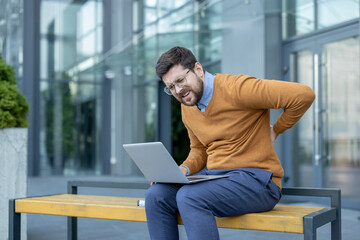 A tired young man sits on a bench near a work building and holds his hand behind his back, feeling...