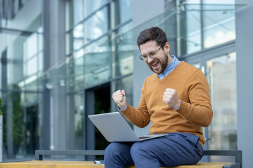 A happy young man is rejoicing, sitting on a bench on the street, looking at the screen of a laptop...