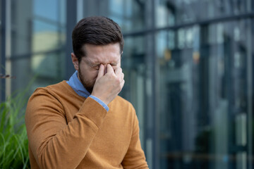 Close-up photo of a young tired and upset man standing outside, rubbing his eyes with his hand