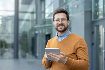 Close-up portrait of a young smiling man in glasses standing outside, holding a tablet and smiling...