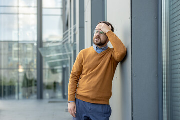 A young tired man stands on the street, leaning against a building and holding his head with his...