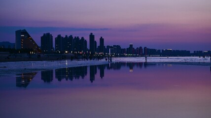 Modern city skyline at dusk reflected on water. A stunning cityscape at dusk featuring tall buildings and their reflections on calm water, with purple and pink hues dominating the sky.
