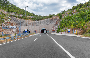 A modern highway approaches the entrance of a road tunnel, flanked by green, rocky mountains under...