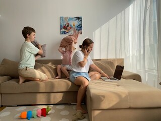 Mom works from home on a laptop while her kids have a pillow fight on the couch. Real-life chaos of...