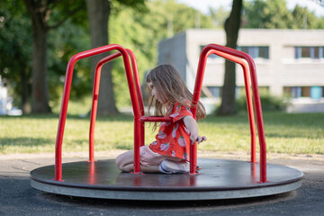 A five-year-old girl rides a carousel on a playground. A Caucasian girl in a red T-shirt walks on...