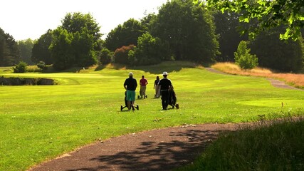 Golfers walking across a green golf course fairway in sunlight, pushing club trolley caddies. Active outdoor lifestyle and leisure sports scene with natural landscape. 