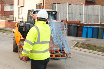 Two construction workers direct a forklift transporting materials through a residential street on a clear day, facilitating ongoing building work © Iryna