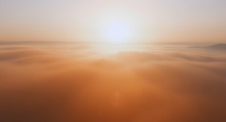 Aerial view of a sunrise above the clouds with golden and orange hues. The sun is partially obscured by the fog. Distant buildings silhouette the horizon.