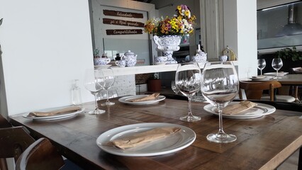Elegant dining table setup in a rustic restaurant. Wooden table with wine glasses, plates, and cutlery prepared for a celebration or special occasion. Beautiful flower bouquet in the background.