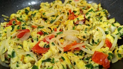 Close-up of homemade scrambled eggs cooking in a black pan. The dish is filled with fresh ingredients like diced red tomatoes, sliced white onions, and chopped green chives. A healthy breakfast.