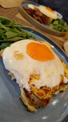 Gourmet breakfast plate featuring a perfect sunny-side-up egg over a savory base with melted cheese and a side of fresh arugula salad. Served on a stylish blue plate in a restaurant.