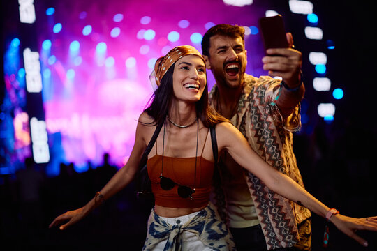 Cheerful couple using cell phone while taking selfie on summer music festival at night.