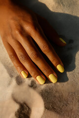Close-up of a hand with butter yellow nail polish resting on sandy surface, captured in warm natural light.

