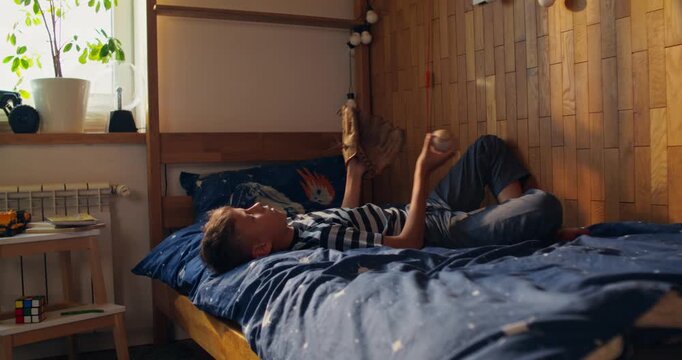 A boy is playing with a baseball, catching it with a glove. He's lying on the bed in his room.