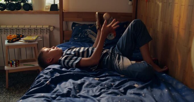 A boy is playing with a baseball, catching it with a glove. He's lying on the bed in his room.