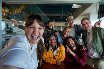 Caucasian woman next to diverse businesspeople smiling and taking a selfie together in a bright, modern office, showing on social media teamwork and camaraderie between work.