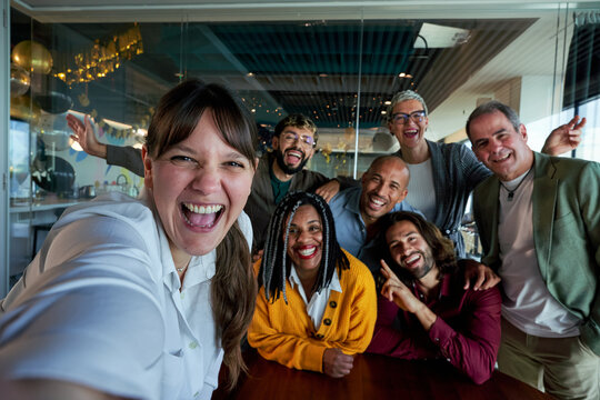 Caucasian woman next to diverse businesspeople smiling and taking a selfie together in a bright, modern office, showing on social media teamwork and camaraderie between work.