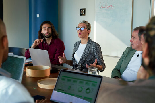 Businesswoman gesturing while leading a team meeting in a modern office, discussing strategy and analyzing data on laptops.