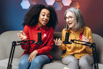 Two women wearing pink ribbon talking into microphones for breast cancer awareness during podcast...