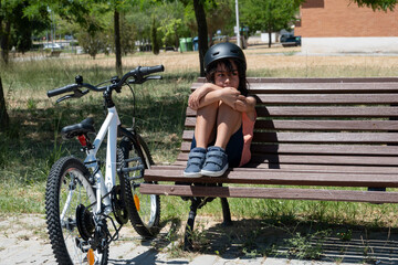 Sad child cyclist wearing helmet sitting on bench in park