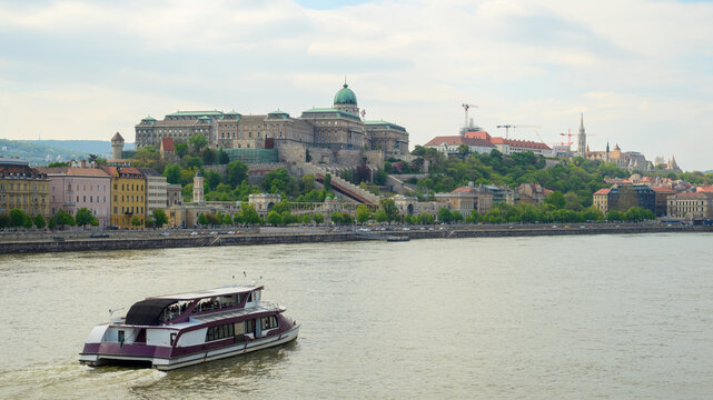 A boat navigates the Danube River with Budapest's Castle Hill in the background. Lush greenery surrounds historic buildings under a partly cloudy sky, creating a picturesque view.