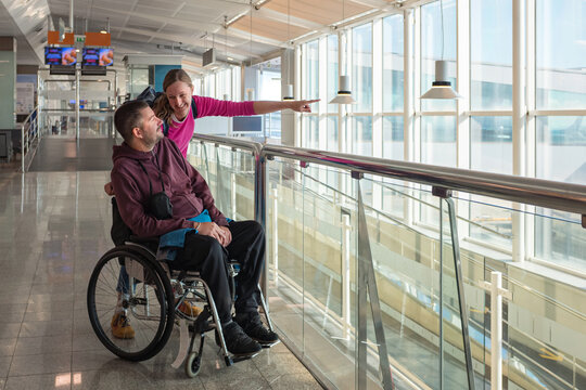 Man with disability using wheelchair with his female personal assistant at the airport. They look out the large windows while preparing for their travel.