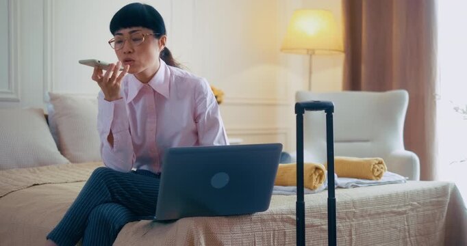 Angry Japanese businesswoman reacting to bad news in a hotel room, reading negative information on her laptop, and recording a voice message on her mobile phone during a business trip.
