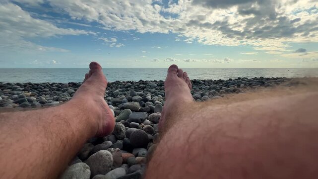 Horizontal 4K travel wide angle video close up. Male hairy bare legs shoes on Black Sea pebble beach. Sun day. Blue cloudy sky. Ocean waves. Concept of summer vacation, tourism, coast line, rest