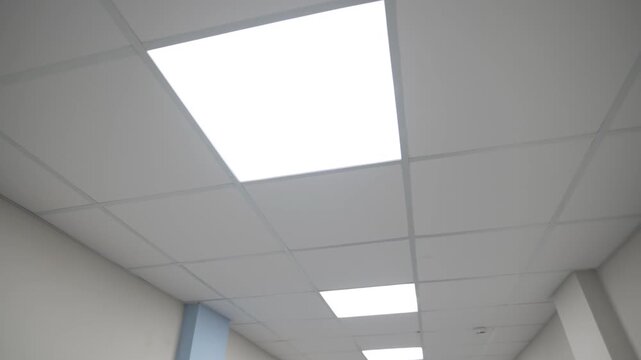 Square led lamps glow on suspended ceiling in hospital corridor. Bright efficient lighting equipment illuminates path in public building