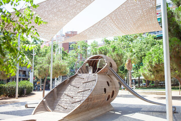 Modern wooden playground structure shaped like a fish under sunshade sails in a green city park on a sunny day
