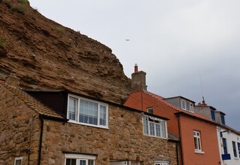 A coastal village scene shows houses nestled against a rock cliff under a cloudy sky  in Staithes -...