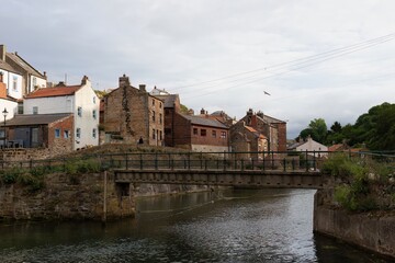 A picturesque view of a town in England. Buildings line a river, with a pedestrian bridge in the foreground  in Staithes - North Yorkshire - United Kingdom