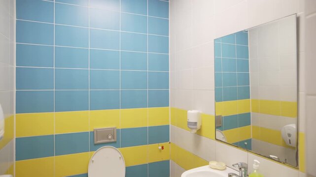 Sink vanity mirror and toilet bowl in restroom decorated with blue and yellow tiles. Washroom illuminated by led light in hospital ward